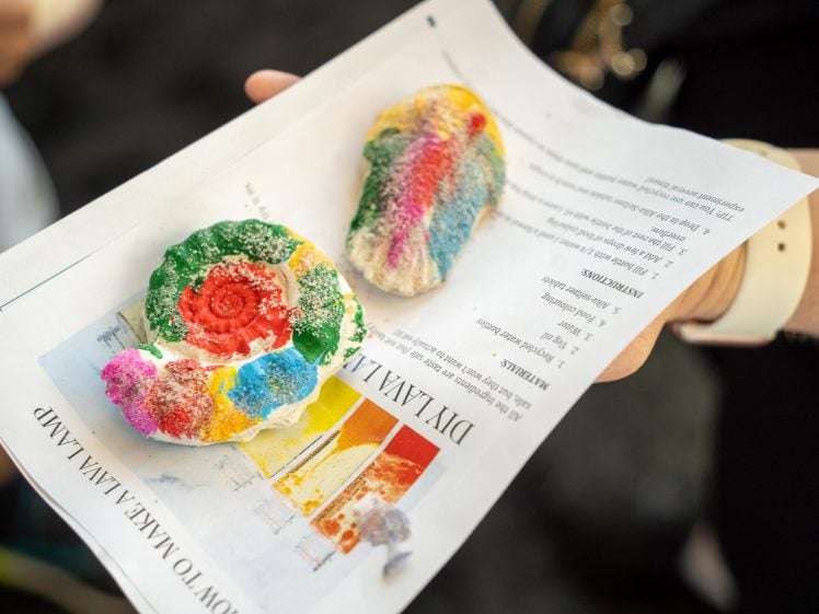 Image of A hand holds a sheet of paper with two colourful, handmade salt dough ornaments covered in rainbow-coloured glitter and paint. The paper has printed text and a small image on it.