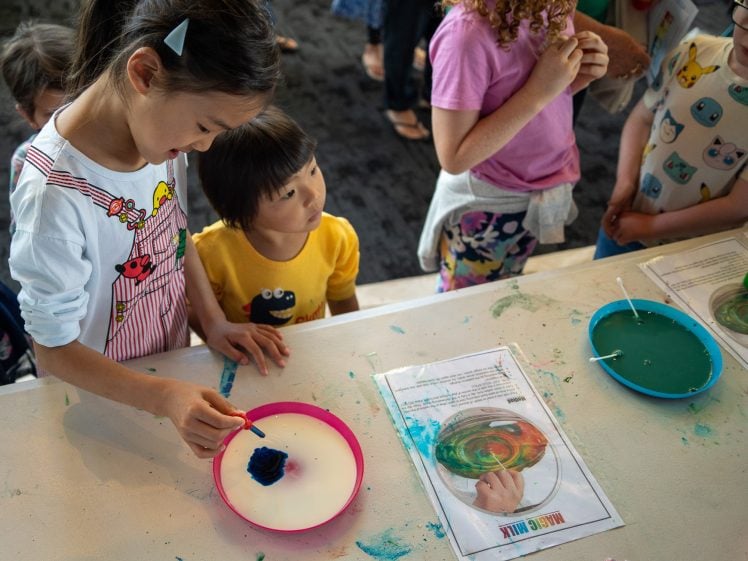 Image of Several children stand around a table doing a colourful milk science experiment with plates of coloured liquid. A girl in striped dungarees holds a cotton bud over a pink plate, while others watch curiously.
