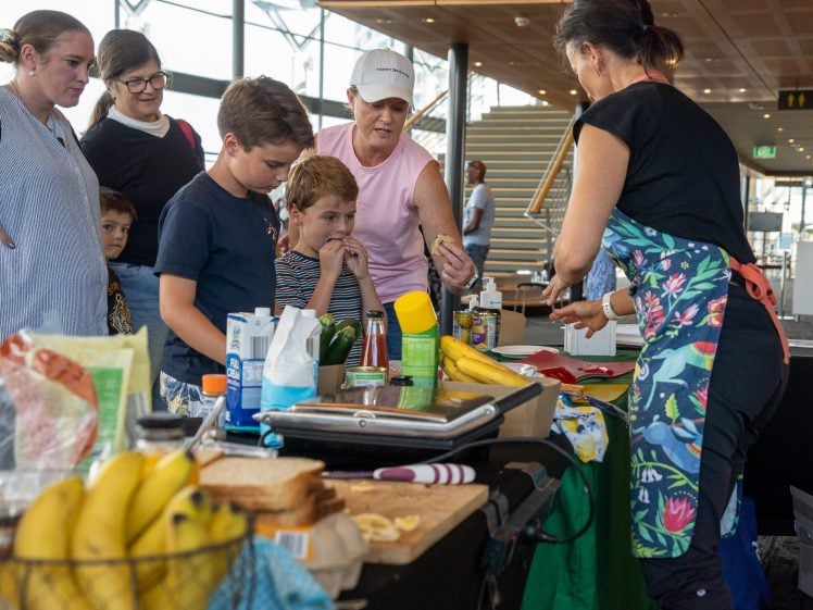Image of A group of children and adults watch a woman in a colourful apron demonstrating food preparation at a table covered with bananas, bread, and cooking supplies in a bright indoor setting.