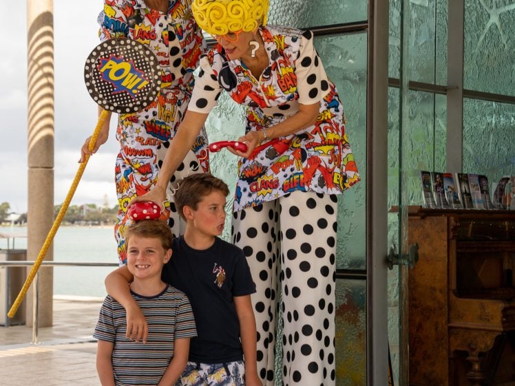 Image of Two children smile for a photo with two performers on stilts wearing colourful wigs, comic-themed outfits, and holding props outside a glass building on a sunny day.