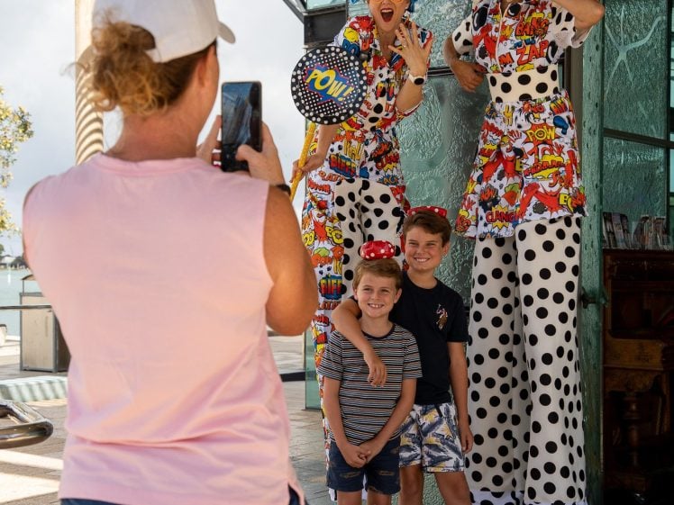 Image of A woman in a pink shirt and shorts takes a photo of two children posing with two performers on stilts wearing colourful, cartoon-themed costumes and large, whimsical wigs outside a modern building.