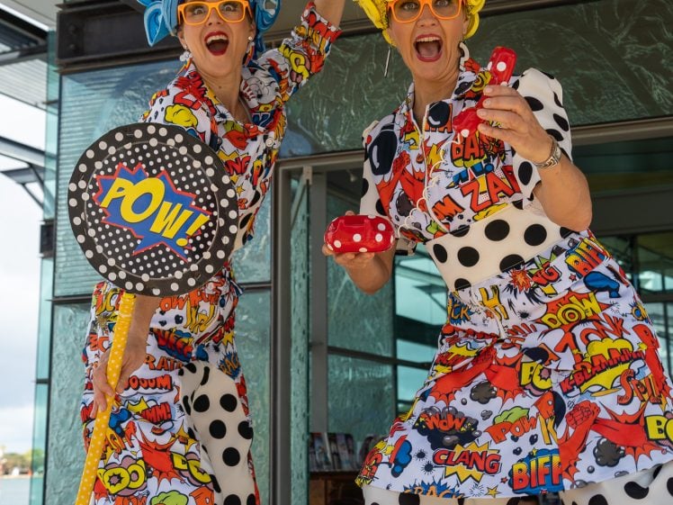 Image of Two people in colourful, comic-book themed costumes and wigs pose energetically. Both wear yellow glasses and hold props. Their outfits feature bold words like “POW!” and “ZAP!” on polka-dot patterns. A child stands in the background.