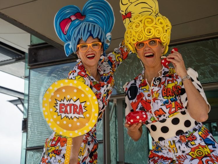 Image of Two people wearing colourful, comic book-themed outfits and oversized, cartoonish wigs smile and pose energetically. One holds a large EXTRA sign, while the other holds a red toy. Both wear bright orange glasses.