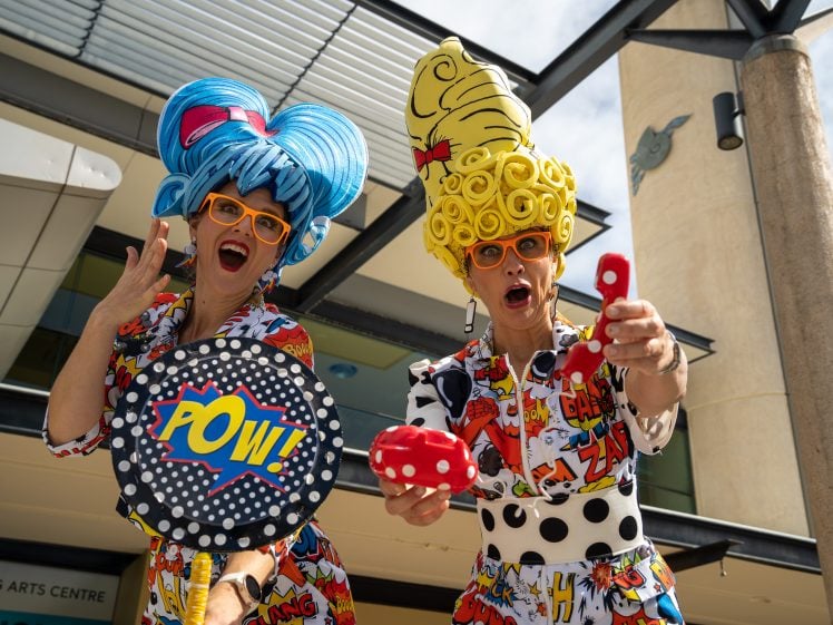 Image of Two women in colourful comic-book themed outfits and wigs pose energetically outside a building. One holds a POW! sign, the other a red prop. Both wear bright orange glasses and expressive facial expressions.