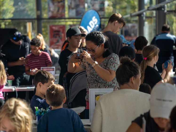 Image of A woman takes a photo with her mobile amid a busy indoor event, surrounded by adults and children exploring various tables and exhibits in a well-lit space.