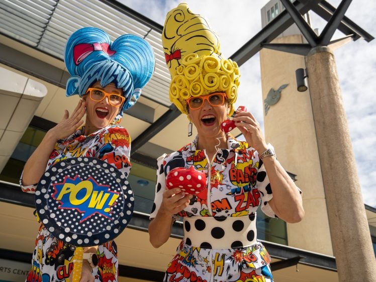 Image of Two people wearing colourful, comic book-themed outfits, giant wigs, and orange glasses pose playfully outside a modern building, one holding a “POW!” sign and the other a toy phone, both smiling enthusiastically.