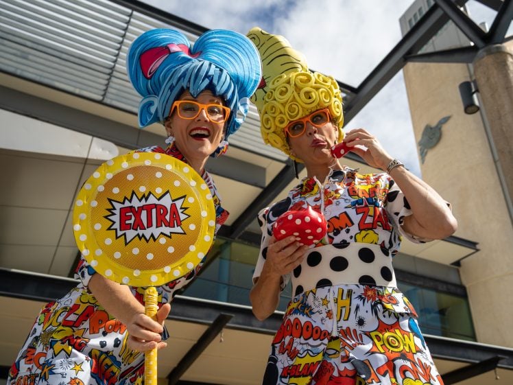Image of Two people in colourful, comic book-themed outfits and oversized wigs pose playfully outdoors. One holds a yellow EXTRA sign, while the other eats from a red polka-dot container. Both wear orange glasses and smile at the camera.
