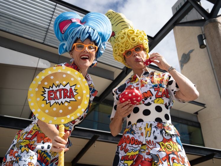 Image of Two people in colourful, comic-themed outfits and exaggerated wigs pose playfully outdoors. One holds a large EXTRA sign, whilst the other eats from a polka-dot cup with a straw. Both wear bright glasses and smile.