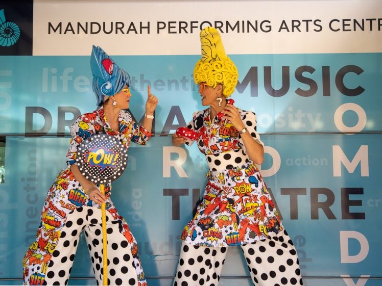 Image of Two performers in colourful comic-themed costumes and large, whimsical hats pose energetically in front of a blue Mandurah Performing Arts Centre backdrop with words like “MUSIC,” “DRAMA,” and “COMEDY”.