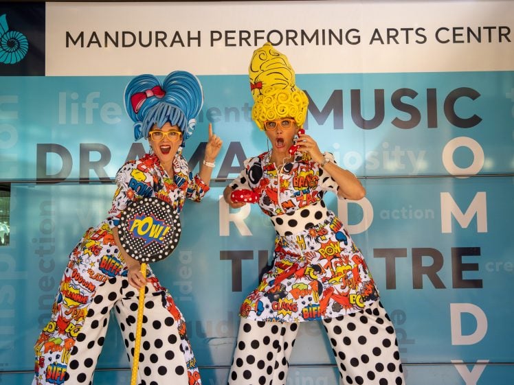 Image of Two performers in colourful, comic-themed costumes and oversized wigs pose energetically in front of a Mandurah Performing Arts Centre sign, holding playful props with animated facial expressions.