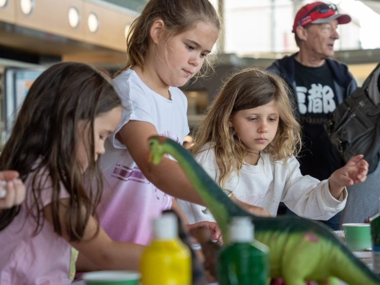 Image of Three young girls focus on a craft activity at a table with paints and a toy dinosaur, whilst an adult in a red cap and glasses stands in the background.