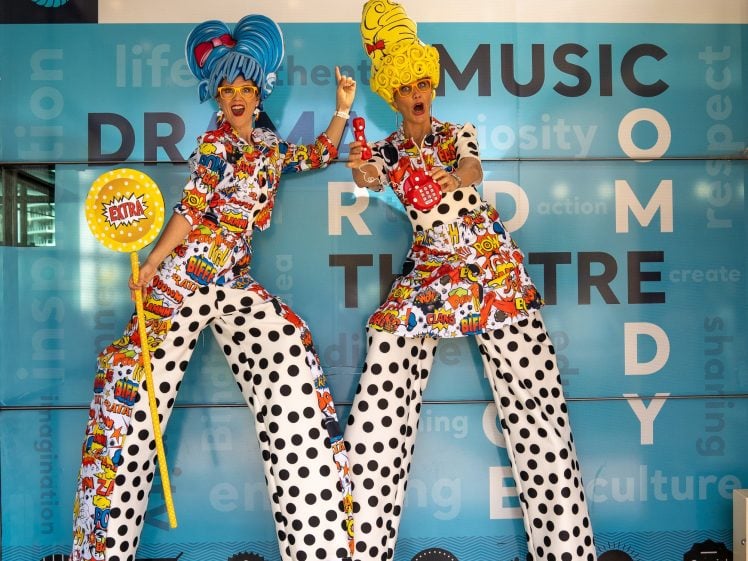 Image of Two performers on stilts with bright blue and yellow wigs wear colourful, comic-themed costumes and polka-dot trousers, standing in front of a blue Mandurah Performing Arts Centre background.
