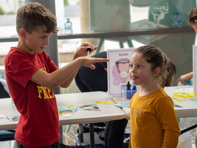Image of A boy in a red shirt holds a dropper over his hand while a girl in a yellow shirt looks up at him, smiling. They are in a classroom with educational taste posters and supplies on the tables behind them.