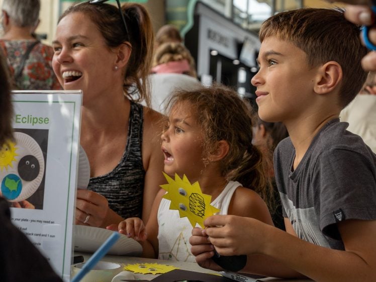 Image of A woman and two children sit at a table, smiling and engaged in a craft activity. The boy holds a yellow sun-shaped cut-out, while the girl looks excited. Other people are visible in the busy background.