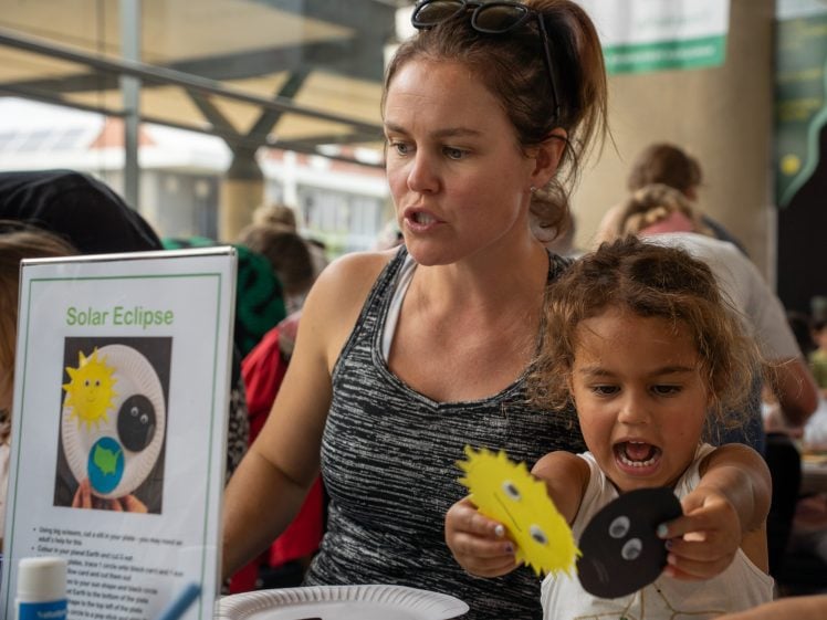Image of A woman and a young girl sit at a table doing a solar eclipse craft. The girl excitedly holds up sun and moon cut-outs. A sign on the table reads “Solar Eclipse” with instructions and a sample craft.