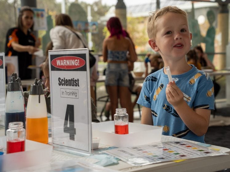Image of A young boy holds a test tube and smiles at a science activity table, surrounded by colourful liquids and a sign reading WARNING! Scientist In Training. People and science displays are visible in the background.