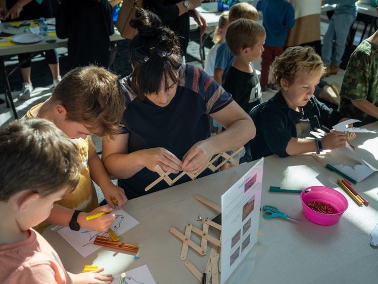 Image of A woman and several children sit at a table crafting with coloured pencils, lolly sticks, and paper in a sunlit room. Supplies are scattered on the table and other people are visible in the background.