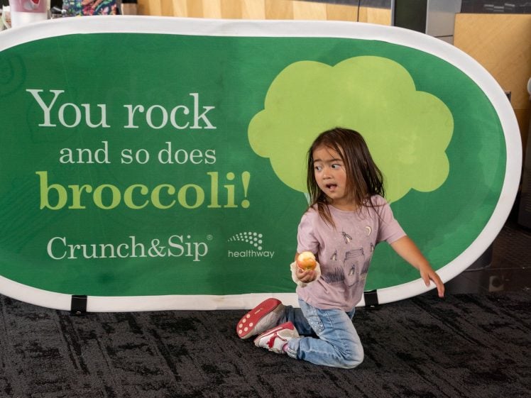 Image of A young girl kneels on a carpet, holding an apple in front of a green sign that reads, You rock and so does broccoli! Crunch&Sip healthway, with a drawing of broccoli in the background.