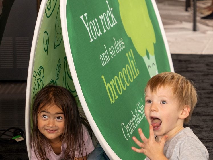 Image of Two young children play under a round green sign with a broccoli graphic that says, “You rock and so does broccoli.” One child smiles whilst the other looks surprised, holding up their hand.