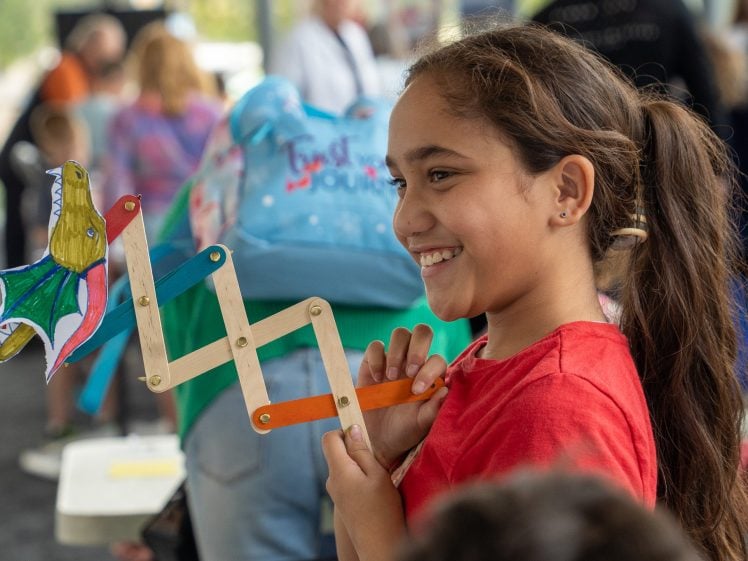 Image of A smiling girl in a red shirt holds a colourful, handmade dragon puppet on sticks at a busy indoor event, with people and tables visible in the background.