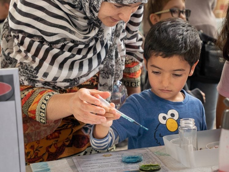 Image of A woman in a patterned headscarf helps a young boy use a dropper to add coloured liquid to trays during a science activity at a busy indoor event. Other adults and children are visible in the background.