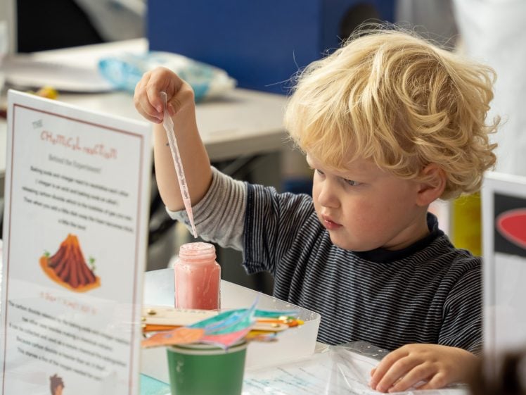 Image of A young child with blond hair uses a dropper to add liquid to a jar with pink substance at a table, engaging in a science experiment or craft activity. Instruction sheets and supplies are visible nearby.