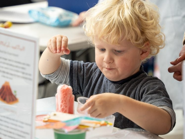 Image of A young child with curly blonde hair is focussed on a science experiment, pouring liquid into a pink volcano model at a table, with informational signs and colourful materials around.