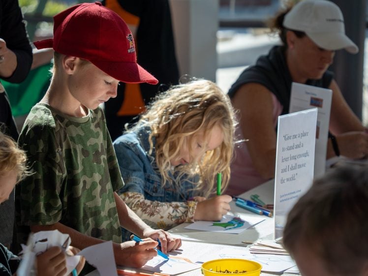 Image of Two children focus on drawing and colouring at a table, surrounded by adults and craft supplies. Sunlight streams in through large windows in the background. A sign and a yellow container of felt-tip pens are on the table.
