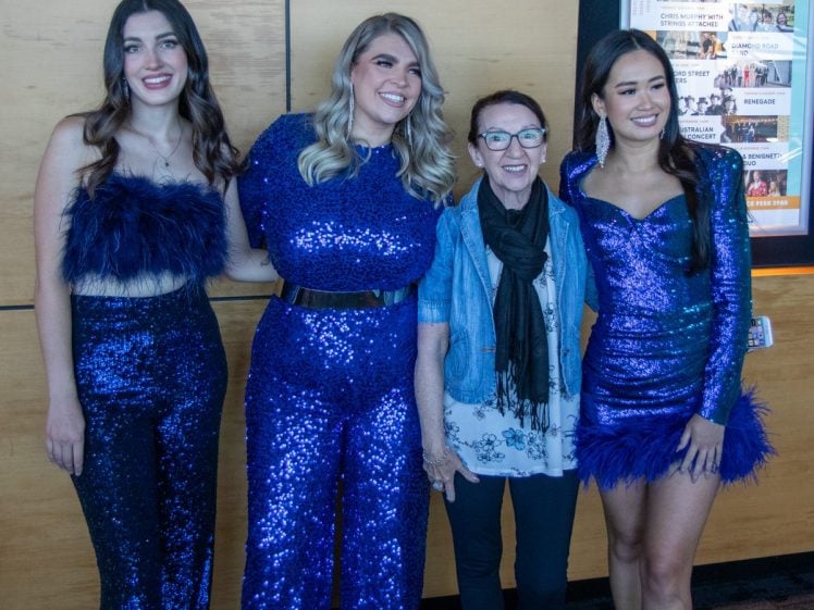 Image of Four women stand together and smile for a photo. Three of them wear matching sparkly blue outfits, while the fourth, older woman wears glasses, a scarf, and a blue jacket. They pose in front of wooden panels and an event poster.