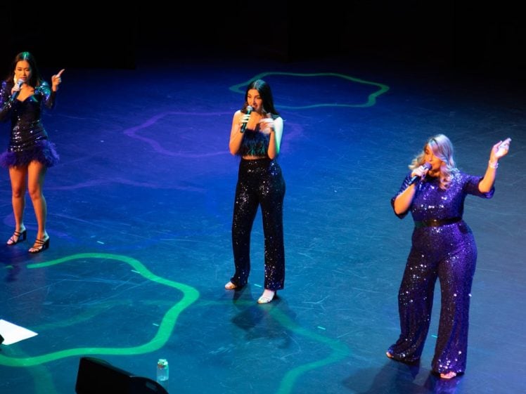 Image of Three women wearing sparkly dark blue outfits sing on a stage with microphones. They are standing apart, each performing to the audience, with colourful patterns and water bottles on the stage floor.