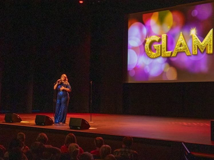 Image of A performer in a sparkling outfit stands on stage with a microphone. On a large screen beside them, the word GLAM appears in gold letters over a colourful, blurred background. The audience is seated in front.