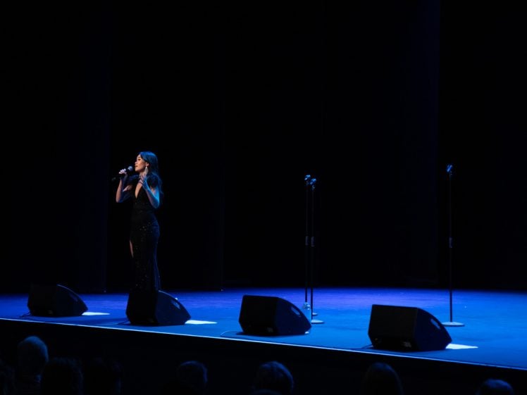 Image of A woman in a black dress sings into a microphone on a dimly lit stage, with three empty microphone stands and blue lighting illuminating the floor.