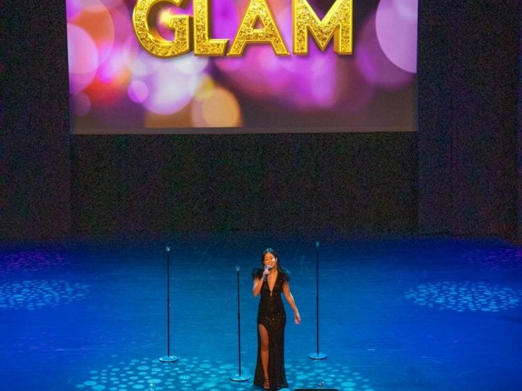 Image of A performer in a black dress sings on stage beneath a large screen displaying the word GLAM in glittery gold letters with colourful, blurred lights in the background.