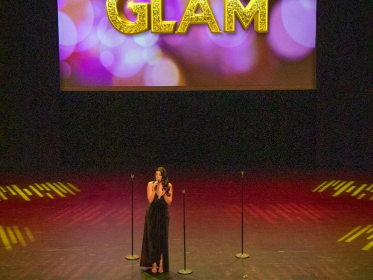 Image of A woman in a long black dress stands on a stage singing, with three microphones around her. Behind her, a large screen displays the word GLAM in glittery gold letters with colourful lights.