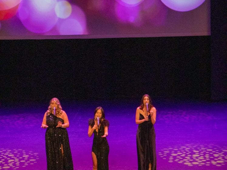 Image of Three women in black dresses sing on stage beneath a large screen displaying the word GLAM in sparkly gold letters with a colourful, bokeh background.