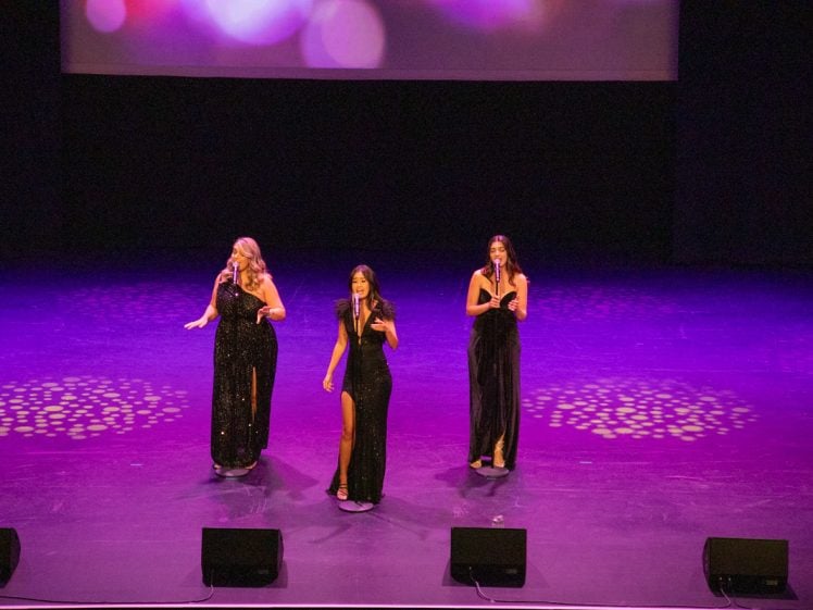 Image of Three women in black evening dresses stand on a purple-lit stage, each holding a microphone and singing. Circular light patterns are projected on the floor, and a screen with blurred lights is visible in the background.