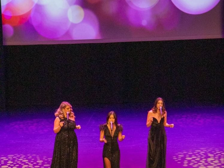 Image of Three women in elegant black dresses sing on stage beneath a large, sparkling GLAM sign, with colourful, blurred lights decorating the backdrop.