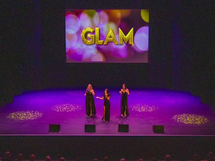 Image of Three performers in black dresses sing on a purple-lit stage, with GLAM in large glittery letters displayed on a colourful screen behind them. Spotlights create circular patterns on the stage floor.