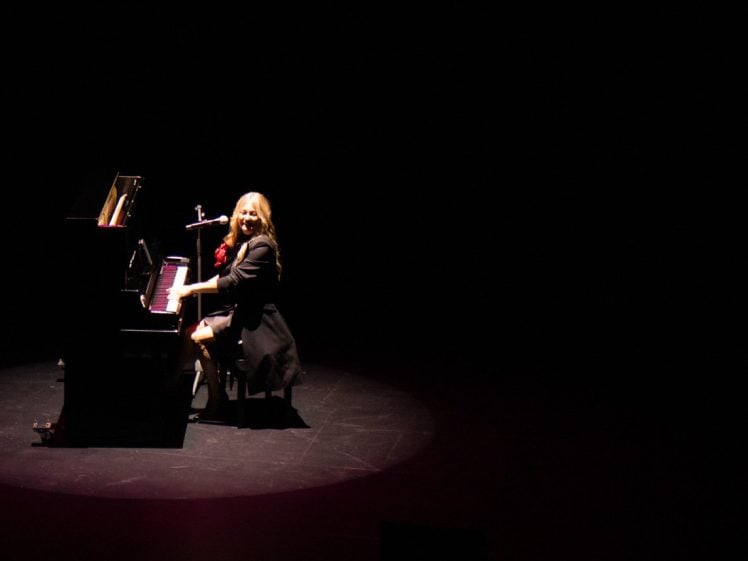 Image of A woman with long blonde hair plays a grand piano and smiles under a spotlight on a dark stage, with a microphone positioned near her.