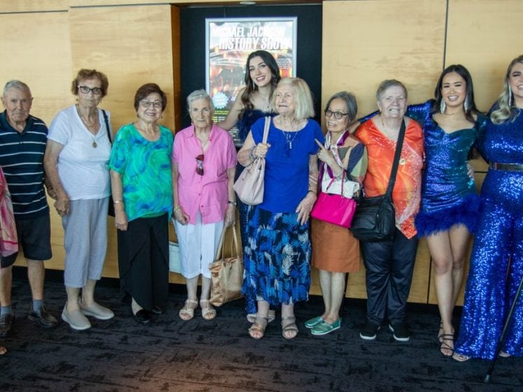 Image of A group of older adults and three younger women in sparkly blue dresses smile for a group photo in front of a beige wall with posters, posing indoors and appearing happy together.
