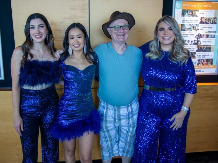 Image of Four people pose together indoors. Three women wear sparkly blue outfits, and a man in the centre wears a blue shirt, checked shorts, and a brown hat. They are all smiling, standing in front of a light wood wall with posters.