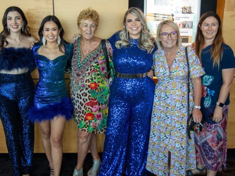 Image of Six women stand together smiling; three wear sparkling blue outfits, two wear floral dresses, and one wears a patterned dress with flowers. They are posing indoors in front of a wooden wall.