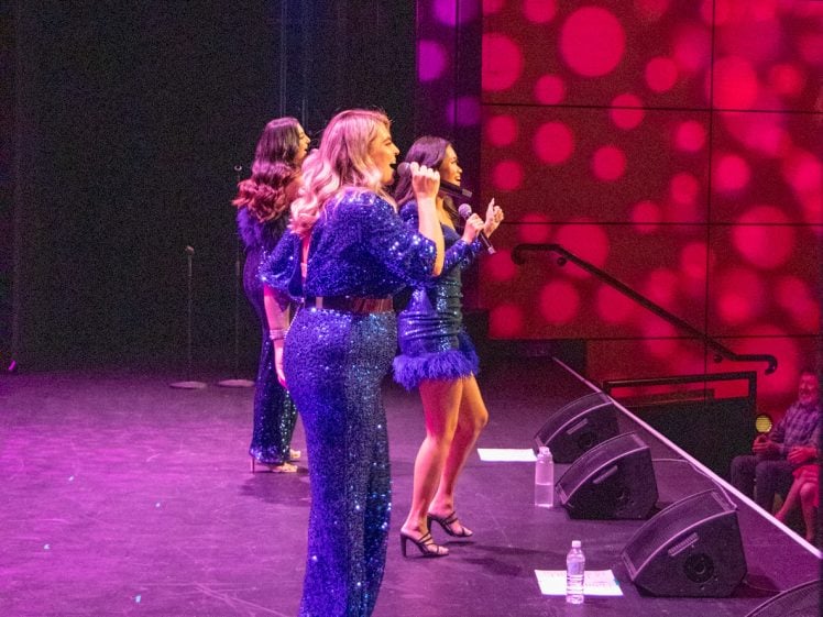 Image of Three women in sparkling blue outfits sing on stage with microphones. Pink circular lights are projected on the wall behind them. Water bottles are on the floor near the performers. An audience is visible in the background.