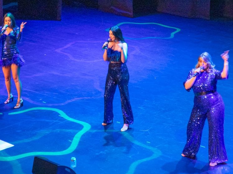 Image of Three women in matching sparkly dark blue outfits sing onstage under blue lighting. Each is holding a microphone, and water bottles are visible on the floor near them.