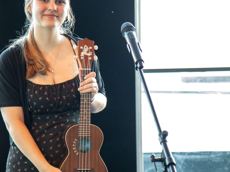 Image of A young woman in a black patterned dress stands indoors, holding a ukulele and smiling softly. She is next to a microphone stand, with light streaming in from a large window behind her.