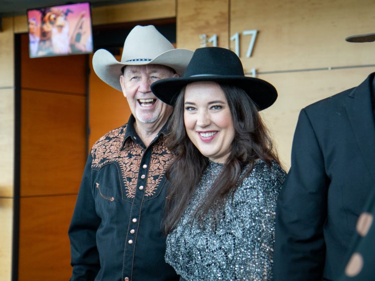 Image of A man and woman dressed in western-style clothing and hats smile at the camera while standing indoors at an event. Another person in a cowboy hat is partially visible beside them.
