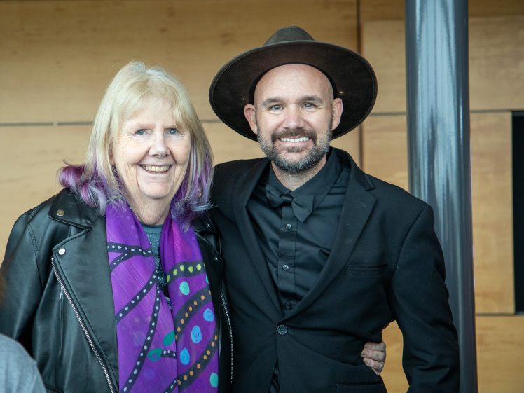 Image of An older woman with blonde and purple hair and a colourful scarf stands smiling next to a man in a black suit and wide-brimmed hat. Both are smiling and posing indoors against a light wood background.