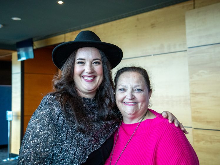 Image of Two women stand closely together and smile at the camera. One wears a black hat and sparkly top; the other has her hair tied back and wears a bright pink top. They are indoors with a wooden wall in the background.