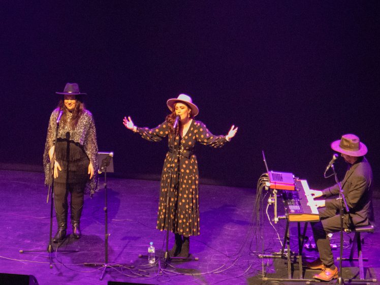 Image of Three musicians perform on a dimly lit stage: two women stand and sing into microphones whilst wearing hats, and a man plays keyboard equipment seated at the side, also wearing a hat.