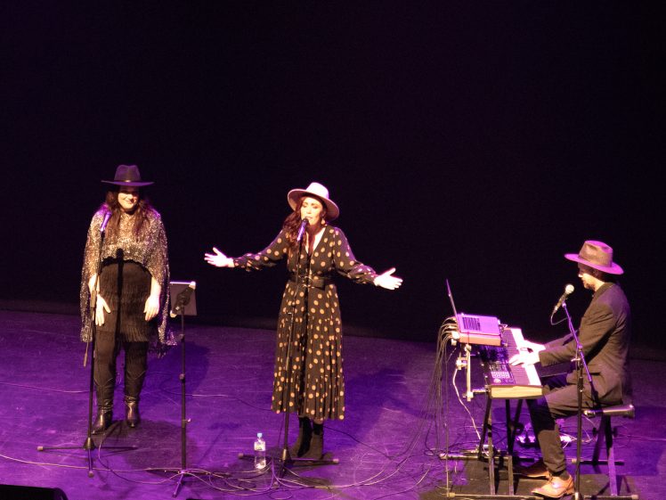 Image of Three musicians perform on stage. Two women, both wearing hats, sing into microphones—one with arms outstretched. A man in a hat plays a keyboard at a table with audio equipment. The background is dark.